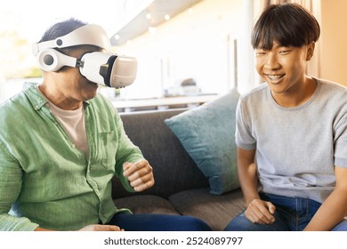 Using VR headset, asian father sitting on couch with smiling teenager at home. Technology, gaming, family, bonding, virtual reality, leisure - Powered by Shutterstock