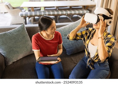 Using VR headset, mother sitting on couch while another mother holding tablet. Technology, virtual reality, digital, interaction, home, leisure - Powered by Shutterstock