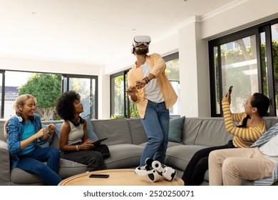 Using VR headset, young man entertaining diverse friends in living room, having fun. Technology, entertainment, virtual reality, socializing, gaming, indoors - Powered by Shutterstock