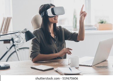 Using VR technologies. Confident young woman in virtual reality headset pointing in the air while sitting at her working place in office - Powered by Shutterstock