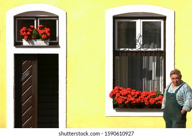 Ustek, Czech Republic, 2005: a man in green overalls, stands proudly in front of his home, with beautifully manicured red geranium flowers on his window sill and matching flowers at the front door - Shutterstock ID 1590610927