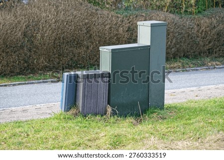Utility boxes and cabinets on a grassy roadside with a hedge background showing infrastructure for electrical and communication services.