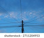 Utility pole supporting a massive, messy tangle of electrical, internet, and communication wires against a bright blue sky with white clouds. A complex web of infrastructure.