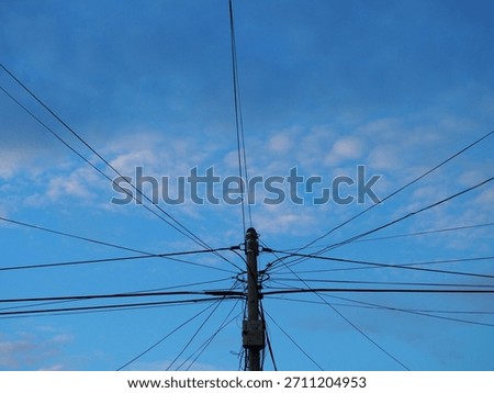 Utility pole supporting a massive, messy tangle of electrical, internet, and communication wires against a bright blue sky with white clouds. A complex web of infrastructure.