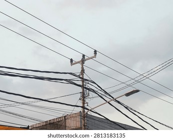 ​A utility pole tangled with a chaotic web of overhead power lines and telecommunication cables against a cloudy overcast sky, featuring a street lamp in an urban or suburban area