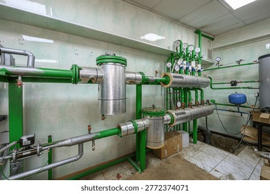A utility room with a complex piping system: insulated metallic pipes, pressure gauges, valves, cylindrical tanks, green pipelines, and a blue expansion tank. Heating water supply setup.