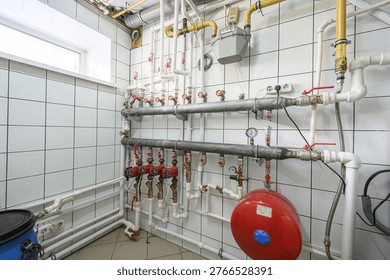 utility room with a complex plumbing system: metal and PVC pipes, pressure gauges, valves, a red expansion tank, gas meter, and tiled walls. Central heating and water supply setup