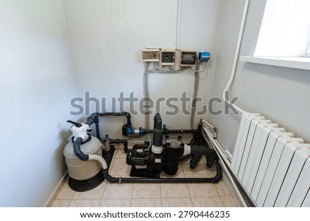Utility room with exposed heating system equipment, including pumps, pressure gauges, and connected metal pipes. A compact control assembly and valves are mounted on the wall near a white heating radiator. Clean tiled floor, bright indoor lighting, and minimal background elements create a clear view of the technical installation
