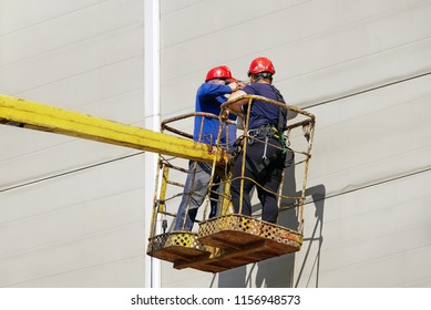 Utility Workers In A Bucket Drill A Wall For Hanging An Outdoor Advertising Sign. Team With Red Hardhat On A Cherry Picker. Professional Working At Height.