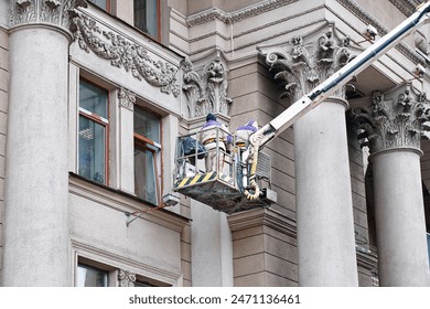 Utility workers on lift platform, facade restoration on historic building with ornate columns, preservation of urban architecture. Men on lift platform clean and maintain exterior of historic building - Powered by Shutterstock