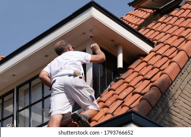 UTRECHT, THE NETHERLANDS - AUGUST 09 2020: Professional House Painter Standing On A Ladder Doing Exterior Paint Work Of A Dormer. Blue Sky Background. Urban City Environment