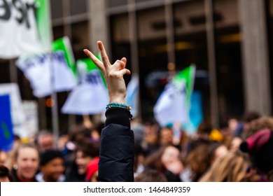 V Gesture At Environmental Protest. A Woman's Arm Is Held In The Air Showing Two Fingers For Peace, As Ecological Activists Are Seen Marching In A City Center.