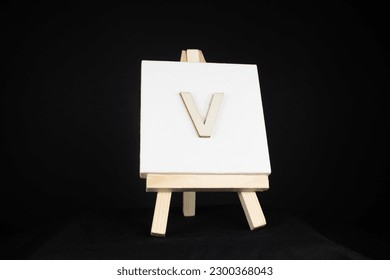 V wooden capital letter and white blank painting canvas resting on an artists easel isolated on a black background