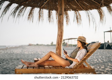 Vacation by sea, weekend on t beach, a beautiful tanned lady in a hat looks at the smartphone screen, sits on a sun lounger on the beach under a straw umbrella. Vacation vacation concept. copy space. - Powered by Shutterstock