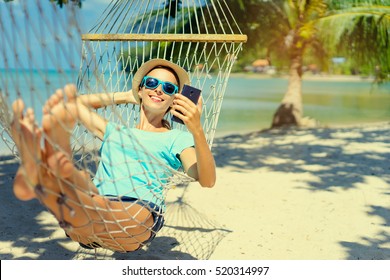 Vacation concept. Enjoying the summer. Young pretty woman in hat and sunglasses using smartphone laying in hammock on the beach. - Powered by Shutterstock