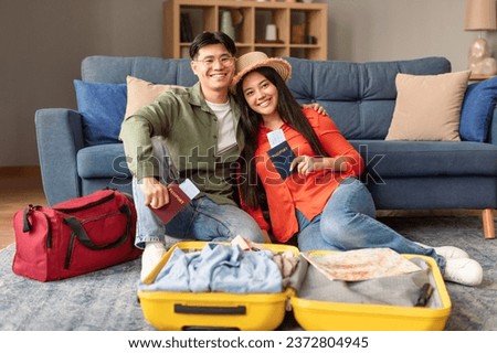 Vacation. Excited asian travelers couple at home, holding travel tickets and passports near an open suitcase, sitting on floor getting ready for vacation travel, smiling to camera