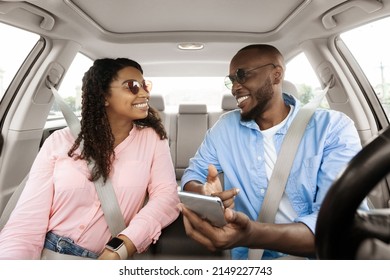 Vacation. Excited black male driver travelling with his beautiful girlfriend on new luxury automobile, showing shortcut way pointing at smartphone screen, couple in sunglasses looking at each other
