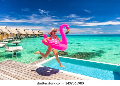Vacation fun woman in bikini with funny inflatable pink flamingo pool float running of joy jumping by infinity swimming pool. Girl enjoying travel holidays at resort luxury overwater bungalow travel.