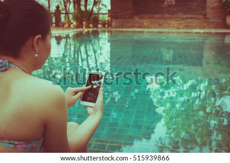 Vacation lifestyle scene of young woman using mobile phone while sitting beside swimming pool in morning time. Weekend and holiday lifestyle with phone addiction concept with vintage filter effect