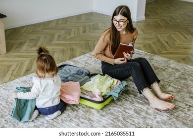 Vacation Preparation. Young Woman Is Packing Suitcase For Trip With Baby. List In Notebook Of Necessary Things.