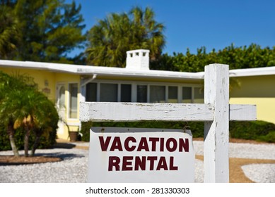 A Vacation Rental Sign In Front Of A Yellow One Story Home On The Beach. 