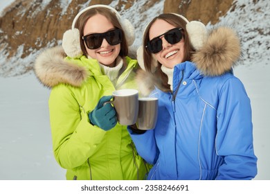 Vacation in a ski resort. Portrait of two fashionable girls in bright downy overalls and trendy sunglasses posing against a snowy winter background. Winter fashion. 