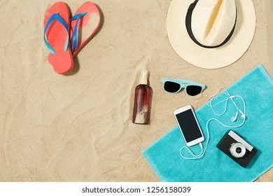 vacation and summer holidays concept - smartphone with earphones and film camera on towel, straw hat, sunglasses, flip flops and bottle of sunscreen oil on beach sand
