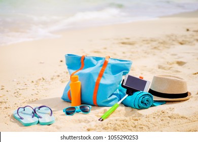 Vacation And Technology. Essentials Accessories On The Sea Beach. Smartphone, Selfie Stick, Bag, Hat, Towel, Sunscreen And Sunglasses.