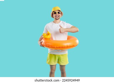Vacation time. Cheerful guy with funny inflatable circle for swimming shows thumb up on light blue background. Joyful skinny young man in shorts, T-shirt and panama recommends summer vacation. - Shutterstock ID 2245924949