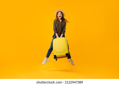 Vacation Travel. Joyful Tourist Woman Jumping In Mid Air Holding Big Suitcase Smiling To Camera Posing Wearing Summer Hat Over Yellow Studio Background. Joy Of Traveling Concept