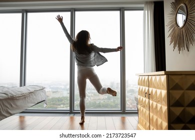 Vacations Of Dream. Back Rear Shot Of Overjoyed Young Female Travelling Abroad Dancing At Large Picture Window Of Luxury Hotel Suite, Happy Woman Houseowner Excited By Modern Flat Apartment Purchased