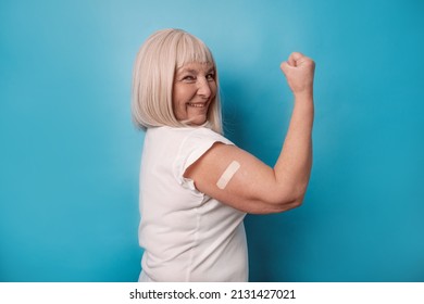 Vaccinated 50s Mature Older Senior Woman Getting A Vaccine Protection The Coronavirus. Happy Female Showing Arm With Bandage After Receiving Vaccination Over Blue Background.
