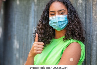 Vaccinated Girl Teenager Teen Mixed Race Biracial African American Female Young Woman Wearing Face Mask In Coronavirus COVID-19 Pandemic Showing Vaccine Band Aid Giving Thumbs Up