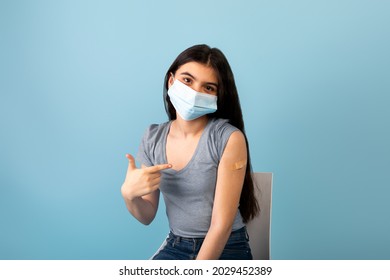 Vaccinated Indian Teen Girl In Medical Mask Pointing At Plaster Bandage On Her Shoulder After Getting Covid-19 Vaccine Shot On Blue Studio Background. Coronavirus Immunization, Health Care Concept