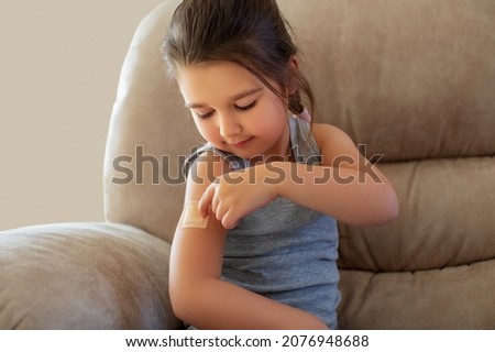 Vaccinated of the little girl is playing at home. Kid with a plaster on his shoulder. Sits on chair Vaccine for covid-19 coronavirus, flu, infectious diseases, Injection, Clinical trials for human.