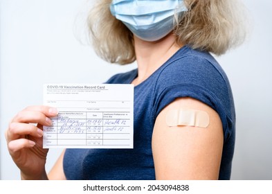 Vaccinated Young Woman In Mask Showing COVID-19 Vaccination Record Card, Healthy Person With Mandate And Plaster On Arm After Getting Corona Virus Vaccine. Proof, People, And Vaccine Shot Concept.