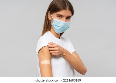 Vaccination Against Covid-19 Outbreak. Portrait Of Serious Millennial Girl In Protective Mask Showing Shoulder With Band Aid After Getting Vaccine Shot On Gray Background