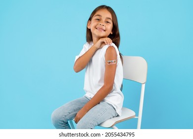 Vaccination Of Children. Happy Vaccinated Kid Girl Showing Arm With Adhesive Bandage After Vaccine Injection Sitting On Blue Studio Background. Kids And Covid-19 Prevention, Antiviral Immunization