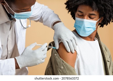 Vaccination Concept. Doctor Giving Shot Or Vaccine Injection To Black Male Patient, Inserting Syringe To His Arm, Young African American Guy In Medical Face Mask Getting Vaccinated Against Covid-19