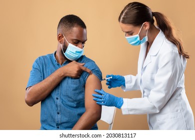 Vaccination And Healthcare During Covid Epidemic. Young Doctor In Face Mask Making Coronavirus Vaccine Injection To Black Male Patient Over Beige Studio Background. Artificial Immunity Concept