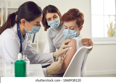 Vaccination And Immunization For Children Concept. Little Child Getting Flu Or Covid-19 Vaccine At The Doctor's. Nurse In Medical Face Mask Giving Injection To Boy Who Came To The Hospital With Mom