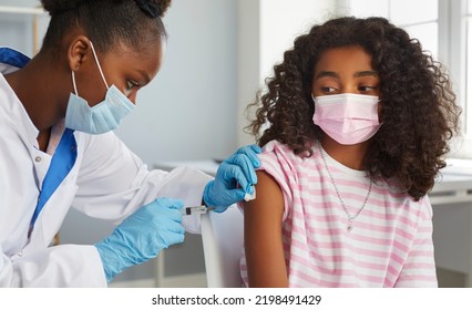 Vaccinations for children. Brave African American teenage girl receives booster dose of coronavirus vaccine. Female nurse in mask and gloves gives injection to child's hand in clinic. Medical banner. - Shutterstock ID 2198491429