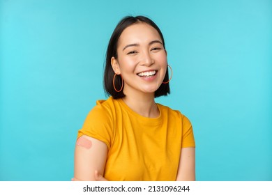 Vaccine campaign from covid-19. Happy and healthy asian girl laughing after vaccination from coronavirus, bandaid on shoulder, wearing yellow t-shirt, blue background