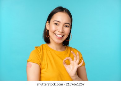 Vaccine Campaign From Covid-19. Happy Healthy Asian Girl Showing Okay Sign And Smiling, Bandaid On Shoulder From Coronavirus Vaccination, Blue Background