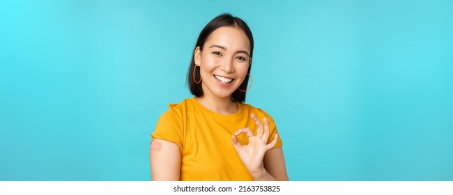 Vaccine Campaign From Covid-19. Happy Healthy Asian Girl Showing Okay Sign And Smiling, Bandaid On Shoulder From Coronavirus Vaccination, Blue Background