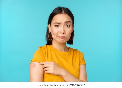 Vaccine Campaign From Covid-19. Sad And Disappointed Asian Woman Showing Shoulder With Bandaid And Grimacing Upset, Standing Over Blue Background