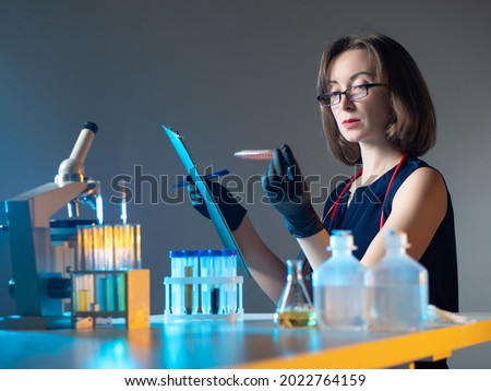 Vaccine development. Woman is developing a vaccine in a lab. She looks at test tube and writes something. Employee of a pharm company creates a vaccine. Test tubes and microscope in front of doctor