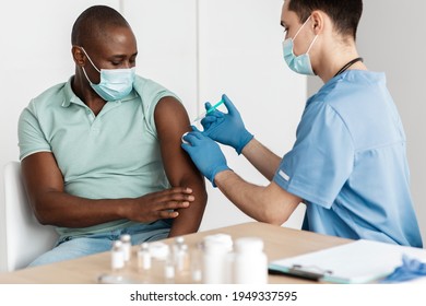 Vaccine, Medicines, Pharmacy And Health Preservation. Young Doctor In Protective Mask Make Injection To Middle Aged African American Patient At Table With Equipment, Vials And Ampoules In Clinic