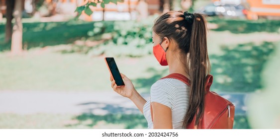 Vaccine Passport COVID-19 App Student Wearing Mask Using Mobile Phone App For Online Class At College Or University. Back To School Woman Holding Cellphone Walking On Campus From Behind Banner.