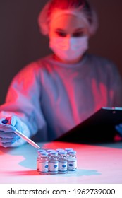 Vaccine Report. Covid-19 Immunization. Medication Record. Female Nurse In Ppe Face Mask Gloves Counting Dose Vials Filling Form On Clipboard In Red Blue Neon Light On Dark Blur Background.